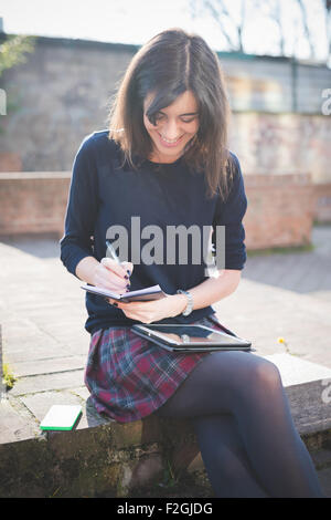 Giovani piuttosto marrone capelli donna in città utilizzando a scrivere su un diario e utilizzando la tavoletta, guardando verso il basso, sorridenti - il multitasking, Foto Stock