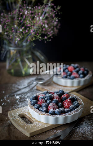 Berry tartetelets con zucchero a velo in piatti di cottura su piastre di legno con bouquet di fiori in background Foto Stock