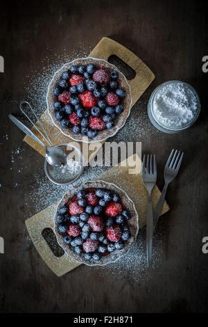 Berry tartetelets con zucchero a velo in piatti di cottura su piastre di legno con il buratto e torta forche. Vista superiore Foto Stock