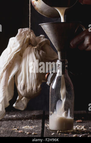 Processo di fabbricazione non a base di latte il latte di mandorle - donna le mani versando il latte dal pan in una bottiglia di vetro attraverso il vecchio imbuto. Vecchia cucina in legno Foto Stock