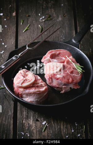 Materie osso buco carni in ghisa pan con carne di vintage forcella, sale, pepe e rosmarino su un vecchio tavolo di legno. Scuro in stile rustico. Foto Stock