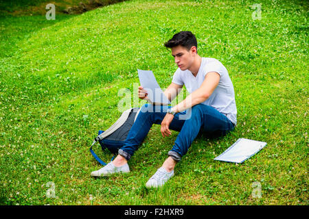 Giovane Maschio Studente le sue lezioni sdraiati sull'erba nel parco cittadino, sorridente in telecamera Foto Stock