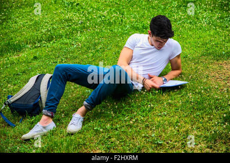 Giovane Maschio Studente le sue lezioni sdraiati sull'erba nel parco cittadino, sorridente in telecamera Foto Stock