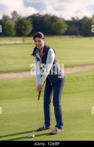 Attraente golfista femminile mettendo sul verde lussureggiante campo da golf, guardando la telecamera con un sorriso sano Foto Stock