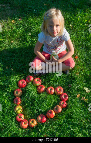 Bambino ragazza bionda con le mele rosse forma di cuore sdraiati sull'erba, per la stagione estiva, frutta, sanitario, alimentare, mangiare, cercando Foto Stock