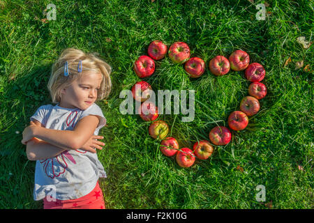 Bambino ragazza bionda con le mele rosse forma di cuore sdraiati sull'erba, frutta, sanitario, alimentare, mangiare, cercando, autunno Foto Stock