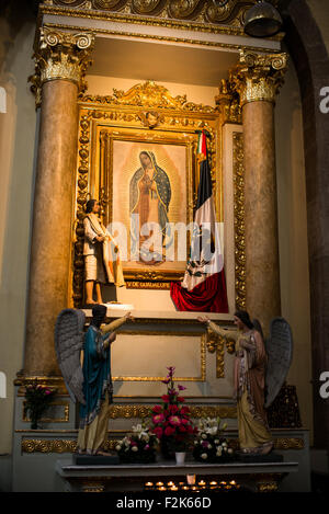 Cappella laterale di Nostra Signora di Guadalupe Sagrario Metropolitano Città del Messico | CITTÀ DEL MESSICO, Messico — Un altare della cappella laterale nel Sagrario Metropolitano presenta una copia dell'immagine di Nostra Signora di Guadalupe, affiancata da una statua di Juan Diego e da due angeli dipinti, con una bandiera messicana drappeggiata accanto — un accoppiamento che riflette il duplice ruolo della Vergine come suprema patrona religiosa del Messico e simbolo di identità nazionale dall'epoca coloniale. L'etichetta "V DE GUADALUPE" è visibile sulla base dell'altare. Foto Stock