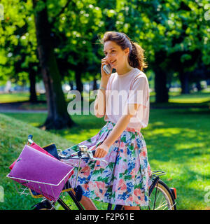 Sorridente ragazza adolescente sul suo bike parlando sul suo telefono cellulare in posizione di parcheggio Foto Stock