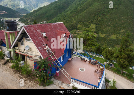 Gli uomini appendere su precariamente per la verniciatura del tetto della casa usando la scaletta di legno in Himalaya vicino Dirang, Arunachal Pradesh, India. Foto Stock