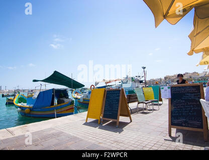 Marsaxlokk ristorante pier con ombrelloni, barche colorate e segni in una giornata di sole, Malta. Foto Stock