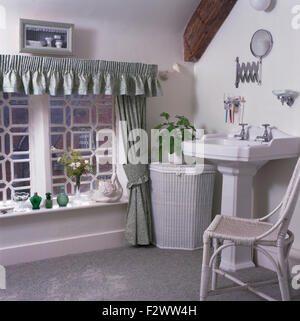Dipinto di bianco vimini cesto per la biancheria e sedia in bianco cottage bagno con verde pallido tende sulla finestra Foto Stock