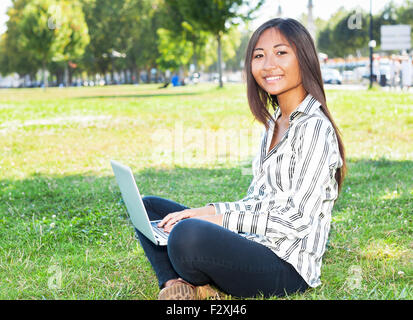 Bella ragazza asiatica su laptop seduto sull'erba Foto Stock