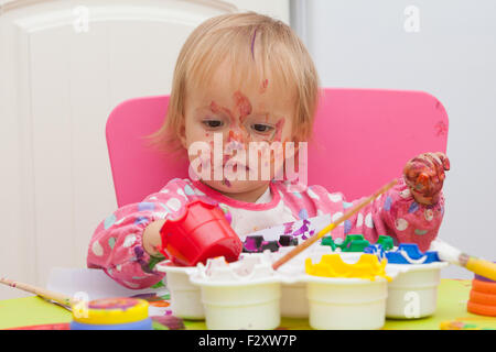 A 16 mesi, capelli biondi, caucasica, ragazza che gioca con vernici e un pennello mentre è seduto dentro a un piccolo tavolo. Foto Stock