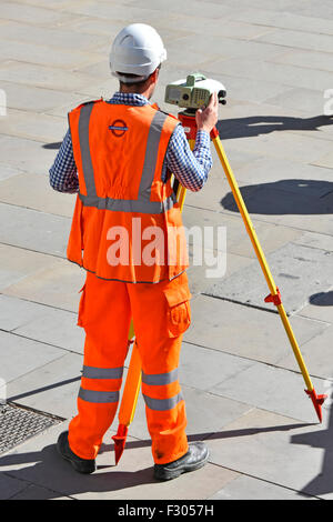 Geometra sul marciapiede pubblico Londra Inghilterra REGNO UNITO utilizzando treppiede montato su apparecchiature di rilevazione indossando alta vis vest contrassegnati sul retro come "underground" Foto Stock