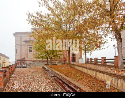 Strada di ciottoli, alberi e la piccola chiesa parrocchiale su sfondo nella nebbiosa mattina autunnale in Piemonte, Italia settentrionale. Foto Stock