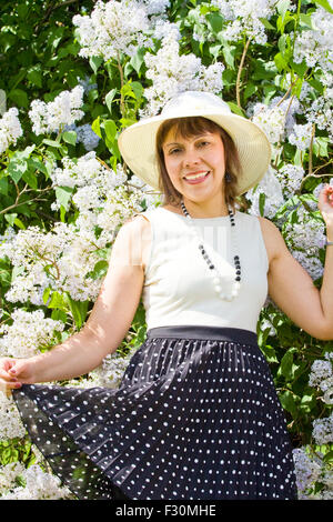 Attraente donna, Europeo, brunette, nel cappello bianco camicia bianca e gonna nera in piedi vicino arbusto di lilla. Foto Stock