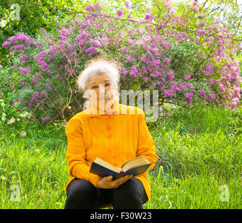Vecchia Signora siede nel giardino e legge un libro di Lilla di arbusti in fiore intorno a. Foto Stock