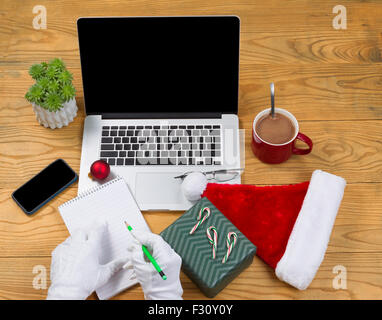 Alta Vista angolato di Santa Claus scrivendo la sua lista di regali con cioccolata calda, computer presente, cappuccio, notepad, matita, telefono cellulare, r Foto Stock