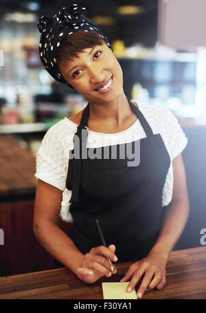 Sincera attraente giovane femmina African American coffee shop proprietario in piedi in un grembiule e bandana dietro il bancone dando t Foto Stock