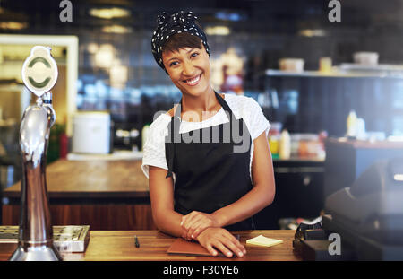 Calda ed accogliente giovani imprenditori in piedi dietro il contatore nel suo cafe dando la telecamera un raggiante sorriso di benvenuto Foto Stock