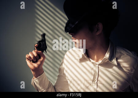 Giovane uomo con la pistola dalla finestra è coperto di ombre dalle persiane Foto Stock