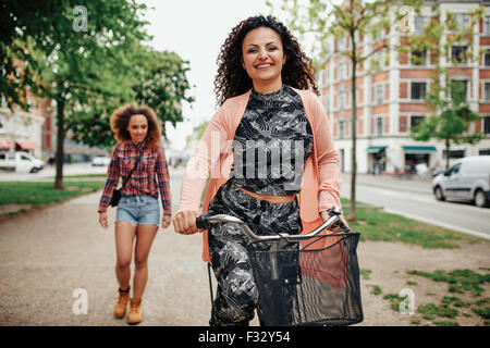 Ritratto di felice giovane donna Bicicletta Equitazione con un altro camminando in background su strada della citta'. Foto Stock