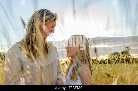 Retroilluminato con un ritratto di una donna (età 25 - 30) con sua figlia (di età compresa tra i 8) sat in in un campo di erba lunga su una sera d'estate Foto Stock