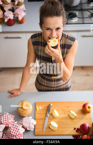 Visto dal di sopra, una donna felice sorrisi come ella è mordere in una di fresco-taglio di apple. Sul bancone della cucina, un tagliere di legno Foto Stock