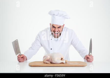 Ritratto del maschio arrabbiato chef cucinare in piedi con coltelli e il pollo sul tavolo isolato su uno sfondo bianco Foto Stock