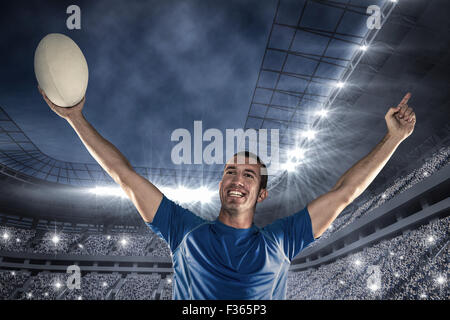 Immagine composita di felice giocatore di rugby in maglia blu tenendo la palla con braccia alzate Foto Stock