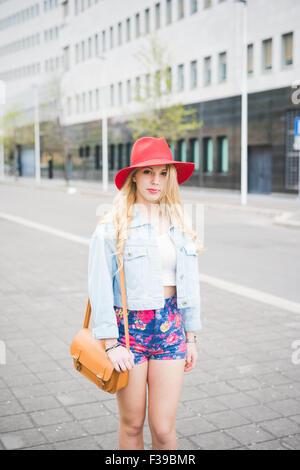 Ginocchio la figura del giovane bella bionda ragazza caucasica in posa la periferia della città vestiti con un Red Hat, un paio di pantaloncini floreali Foto Stock