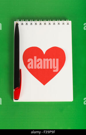 Cuore rosso con una penna nera sul notebook bianco su sfondo verde Foto Stock