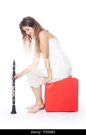 Bruna giovane donna si siede su uno sgabello rosso in abito bianco e trattiene il clarinetto in studio e sorrisi Foto Stock