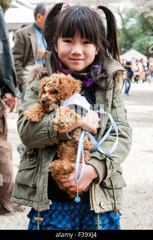 Bambino giapponese, ragazza, 9-10 anni in cappotto, sorridente e creando per il visualizzatore mentre lei coccole e tiene in entrambi i bracci il suo cane. Contatto visivo. Foto Stock