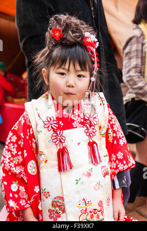 Cinque anni ragazza giapponese, leggermente al di sopra del peso con chubby guance, stando in piedi in posa per viewer indossando rosso e rosa kimono. Cheeky espressione. Foto Stock