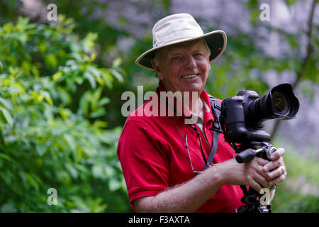 Sorridente uomo anziano che indossa Tilley Hat tenendo la macchina fotografica sorridente a. fotografo sfondo sfocato fogliame Foto Stock