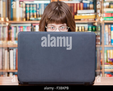 Vista diretta della ragazza con occhiali legge da schermo di computer portatile in biblioteca Foto Stock