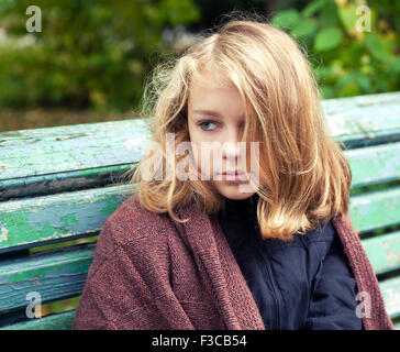 Bella bionda caucasica ragazza adolescente in marrone plaid di lana seduta sul vecchio banco verde nel parco di autunno Foto Stock