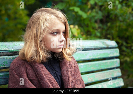 Bella bionda caucasica ragazza adolescente in plaid di lana seduta sul vecchio banco verde nel parco di autunno Foto Stock