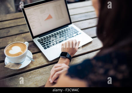 Mano femminile che indossa un smartwatch seduti ad un tavolo con il computer portatile e la tazza di caffè. Donna al lavoro su computer portatile presso il cafe. Foto Stock