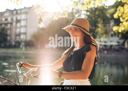 Ritratto di felice giovane donna presso il parco della città passeggiando lungo un laghetto con la sua bicicletta. Unione modello femminile indossando hat cerca