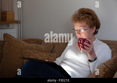 Una donna di mezza età in un momento di relax a casa con una tazza di caffè mentre navigano in Internet utilizzando un tablet ipad computer Foto Stock