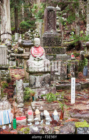 Giappone, Koyasan, Okunoin cimitero. Una linea di jizo bosatsu statue sinistra sulla parete rocciosa come offerta di fronte alcune pietre tombali nel ceder foresta. Foto Stock