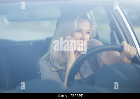 Giovane donna sorridente usando il telefono in un'auto. Bella ragazza bionda in chat smartphone durante la guida Foto Stock