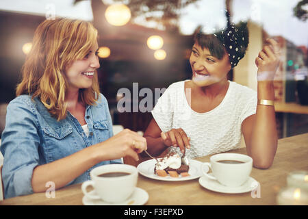 Due piuttosto giovane donna godendo di caffè e una fetta di torta insieme in una casa di caffè seduti ad un tavolo a ridere e spettegolare con felice Foto Stock