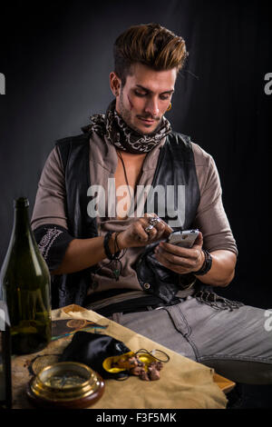 Moda Uomo Vestito da pirata con un telefono cellulare da indossare giubbotto in pelle e sciarpa seduto nella sedia accanto al tavolo, con vuoto Bott Foto Stock