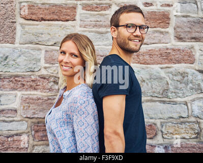 Coppia felice in piedi di schiena e sorridente alla fotocamera contro un muro di mattoni sfondo Foto Stock