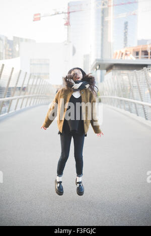 Giovane bella asiatica hipster lunghi marrone capelli dritti donna nella città di saltare sul posto, cercando nella fotocamera, sorridente, durante il tramonto in controluce - carefreeness, concetto della gioventù Foto Stock