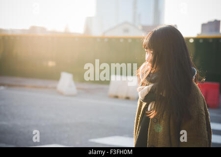 Profilo Ritratto di giovane bella asiatica hipster lunghi marrone capelli dritti donna della città e si affaccia a destra, pensieroso, in città durante la luce del tramonto - riflessivo, pensando di future concept Foto Stock
