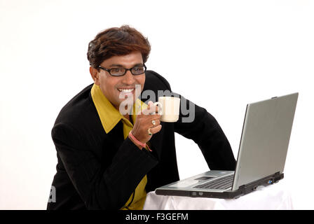 Indian uomo cappotto nero occhiali seduta laptop computer tenendo tazza da caffè signor#364 Foto Stock